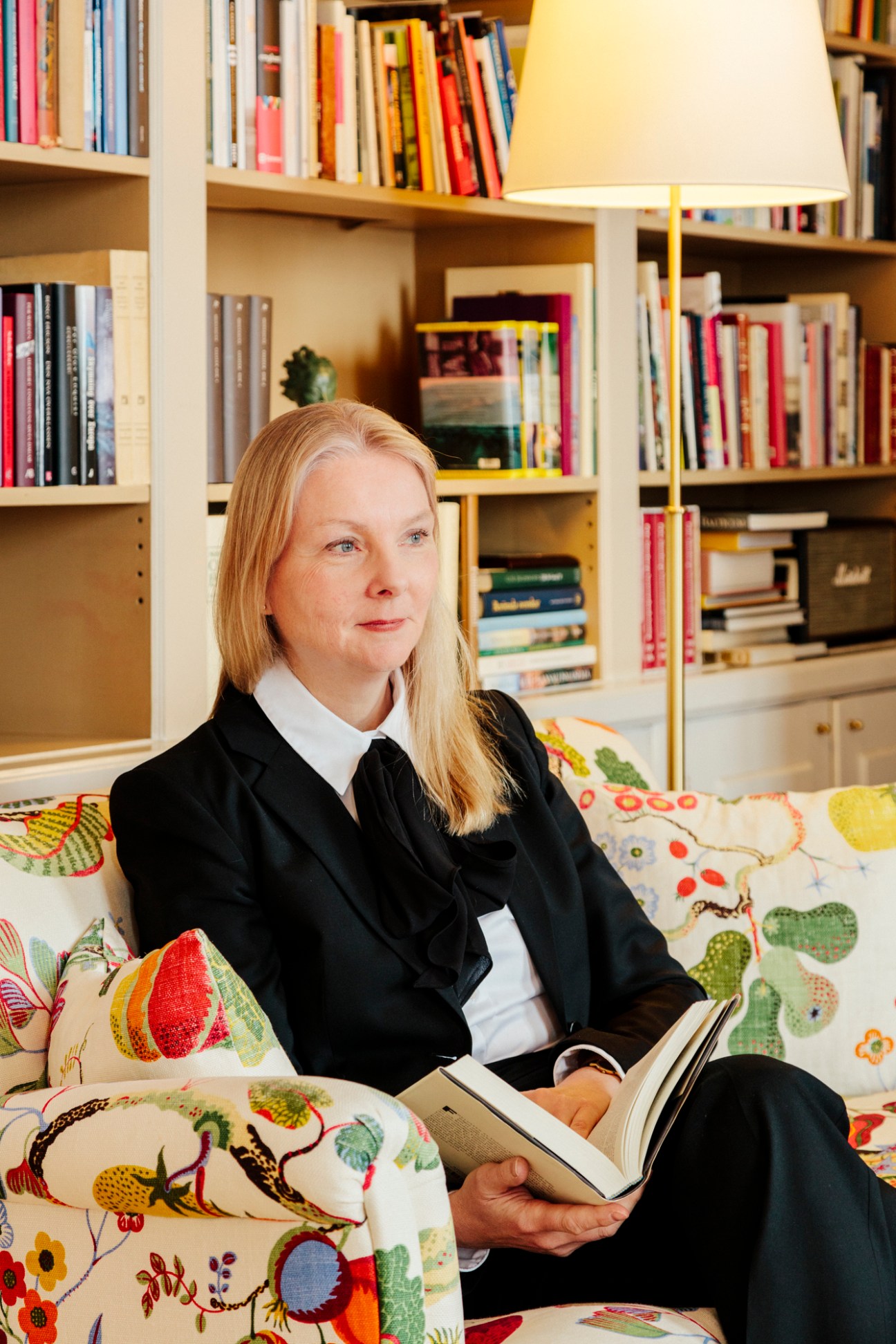 Nicola Clase, Sweden's ambassador to Finland, sitting on a floral sofa reading a book in a room with bookshelves