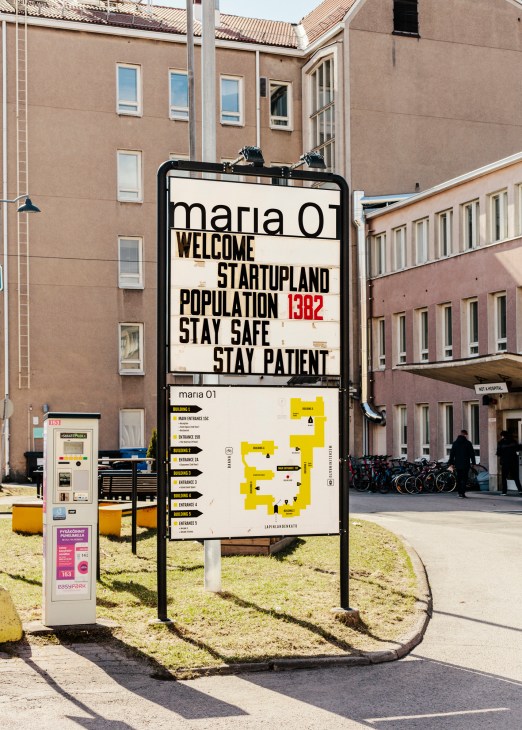 Large exterior sign for Maria 01 displaying "Welcome Startupland Population 1382 Stay Safe Stay Patient" with building directory below.