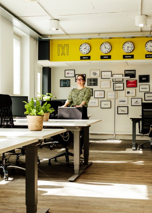 A woman working at a desk in a bright modern office with yellow accent wall displaying world clocks and company logos.