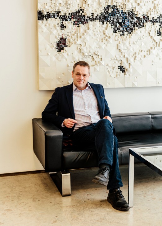 Mayor of Helsinki, Jan Vapaavuori, sitting on a black leather sofa in front of a three-dimensional wall art installation.
