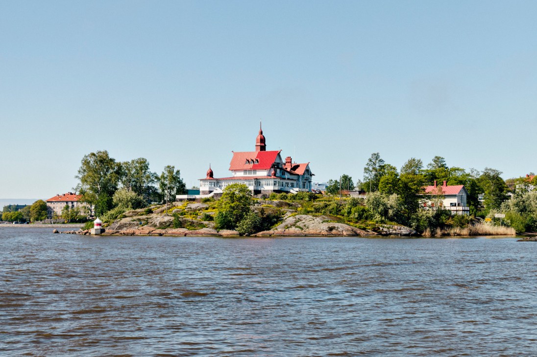 A historic building with a red roof sits on a rocky, tree-covered island surrounded by Baltic Sea waters.