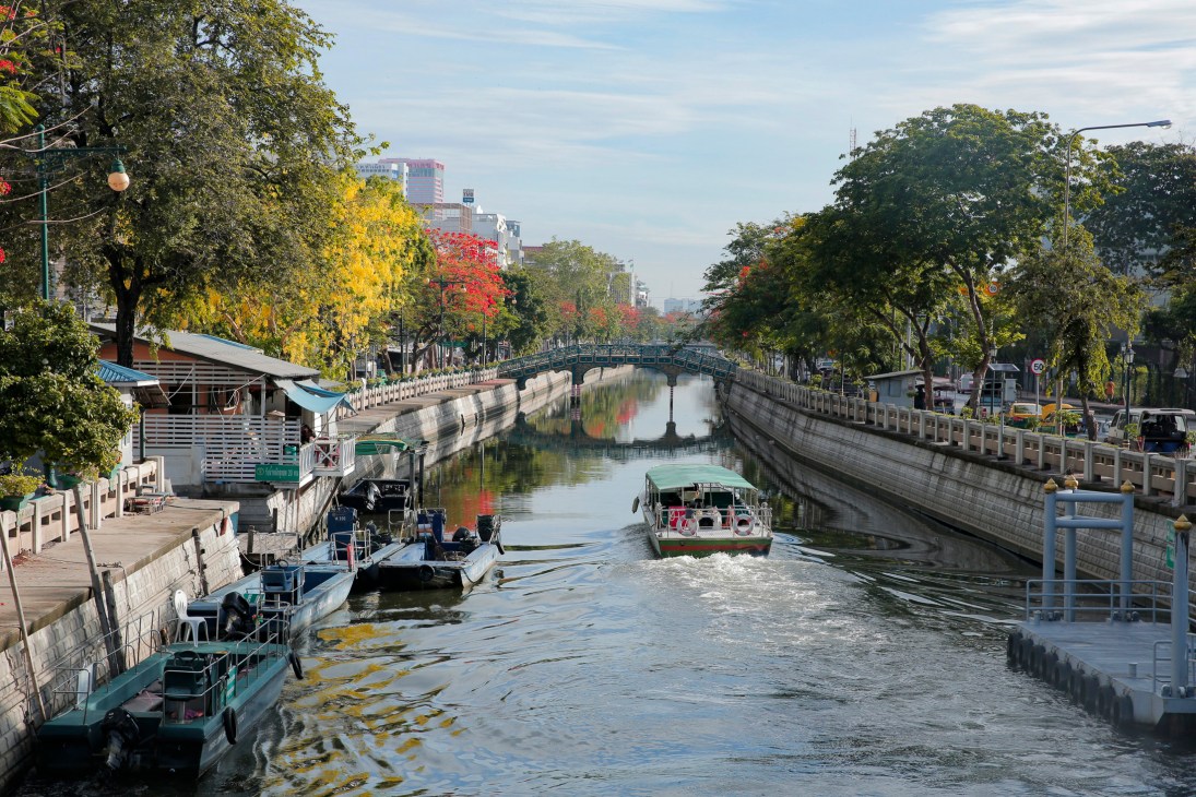 Modern electric ferry boat on Bangkok canal with traditional Thai temple and urban buildings in background