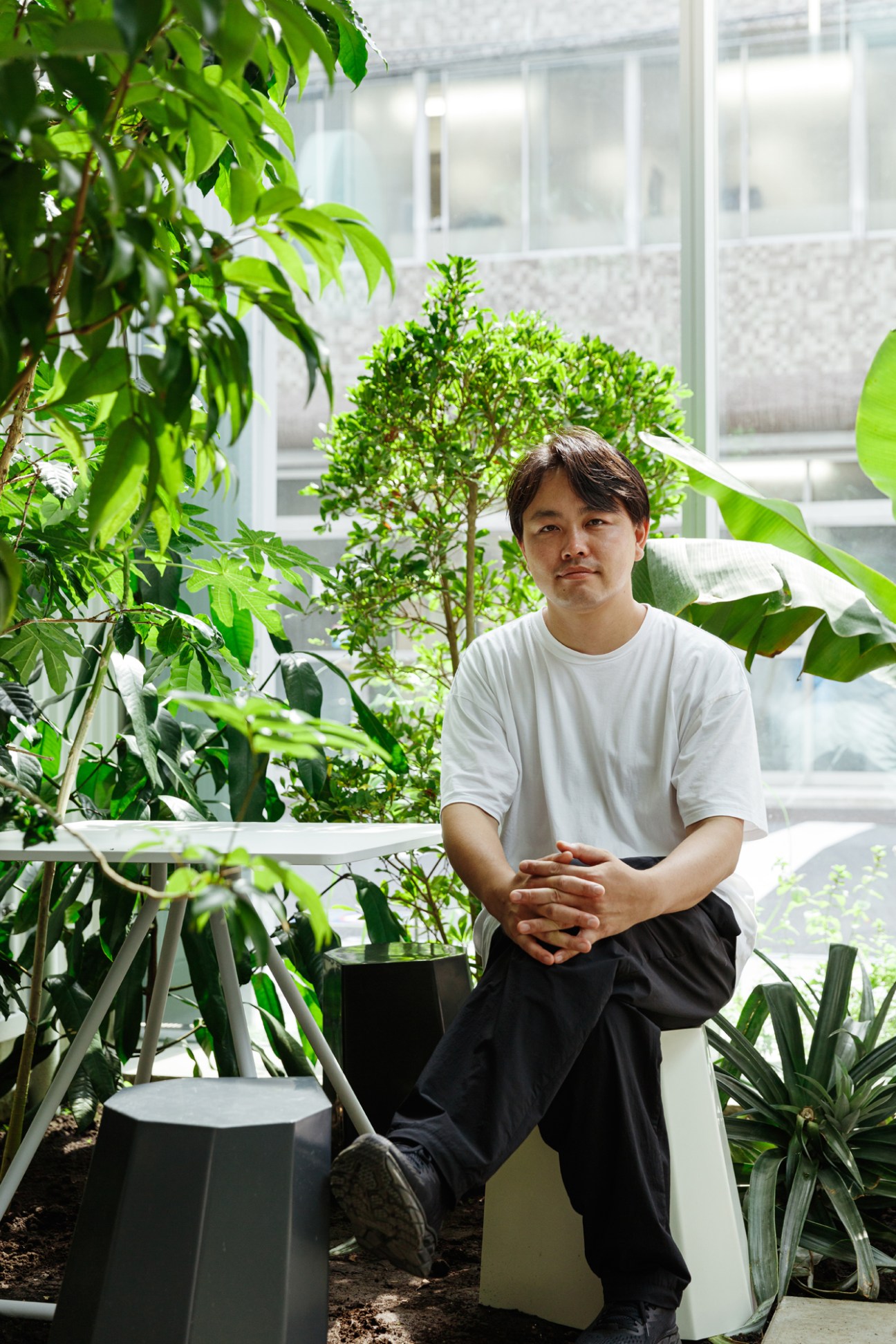Man in white t-shirt seated among lush green plants in a bright office with large windows.