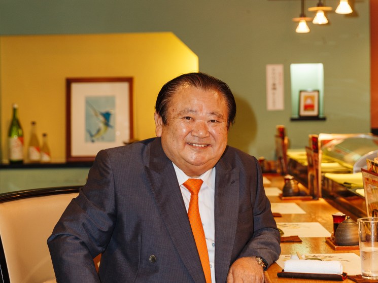 A smiling older man in a grey suit and orange tie seated in what appears to be a Japanese restaurant.