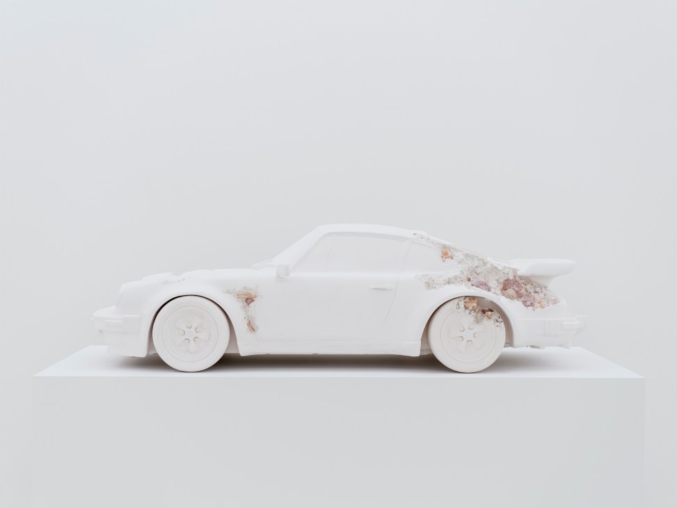 White sculptural car form with weathered patches and circular wheels against minimal background