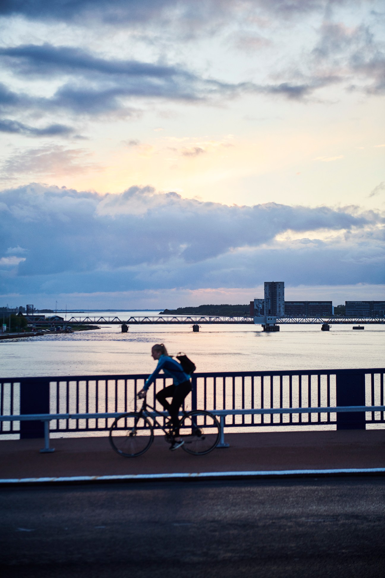 Cyclist riding across Limfjordsbroen bridge in Aalborg with industrial waterfront buildings and cloudy sky in background.