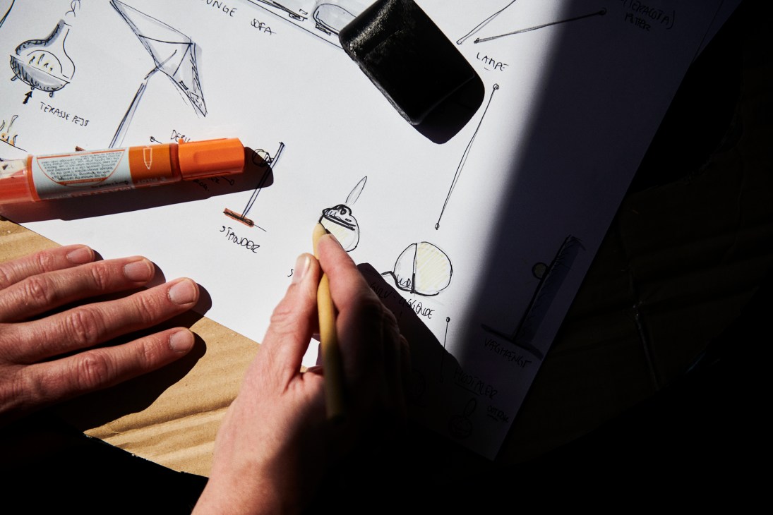 Designer sketching product concepts with orange marker and black container on white paper with hand-drawn notes.