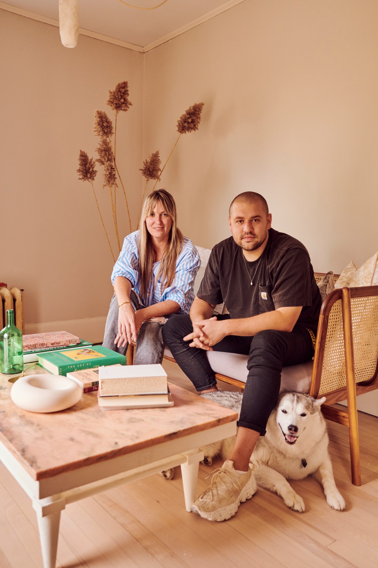 Two people sit on chairs in a minimalist interior with dried flowers and books on a table, alongside a white dog.