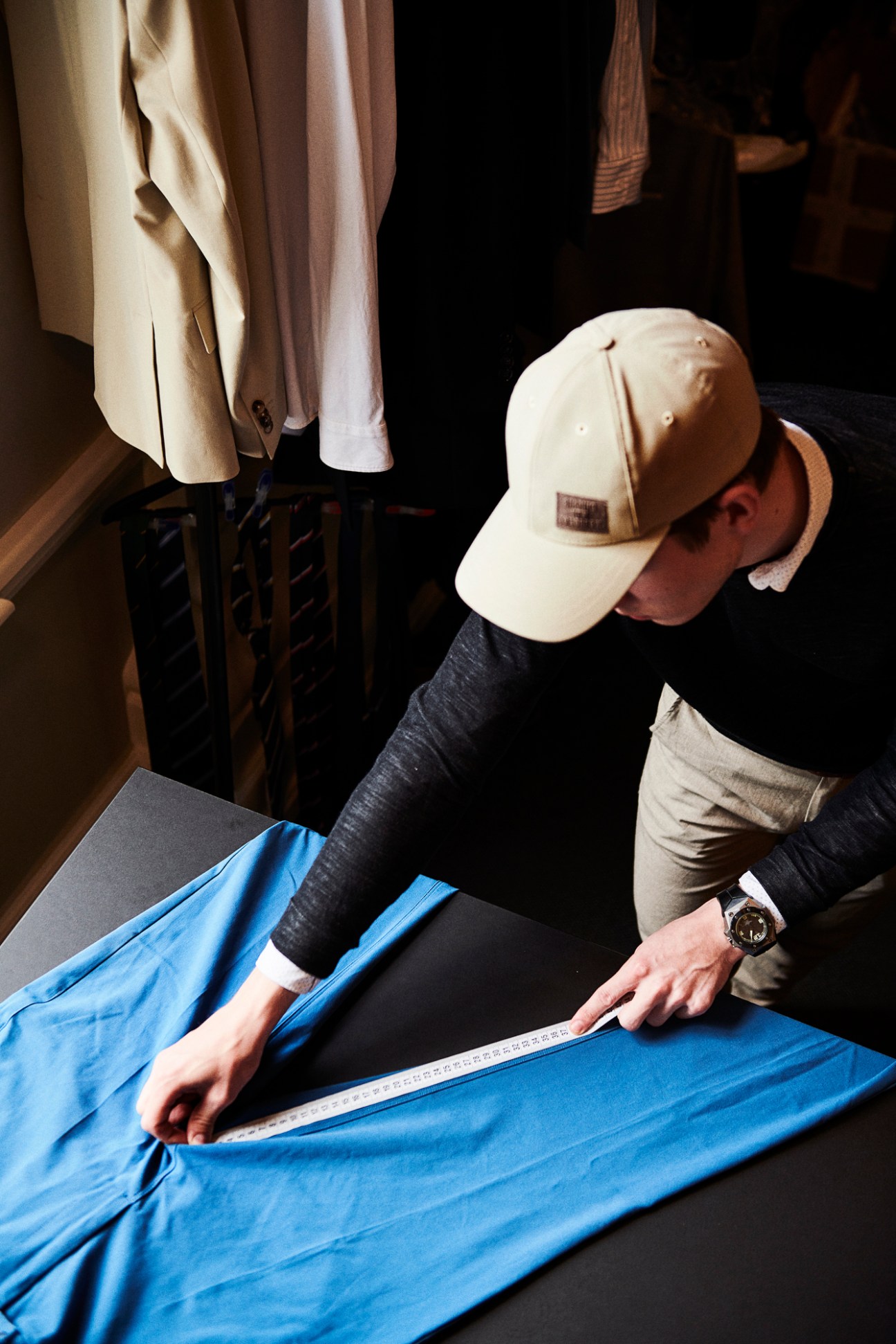 Person in baseball cap measuring blue fabric with ruler on dark table in design workspace with hanging clothes