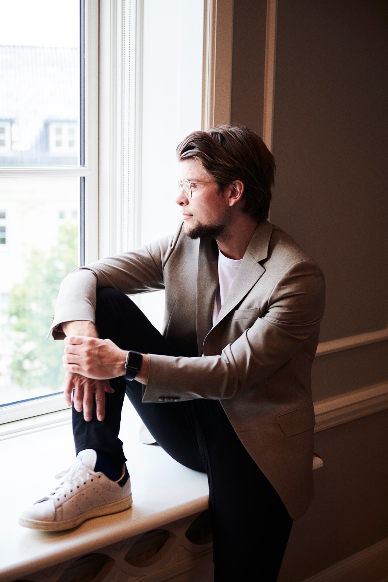 Man in beige blazer sitting by window wearing glasses and smartwatch