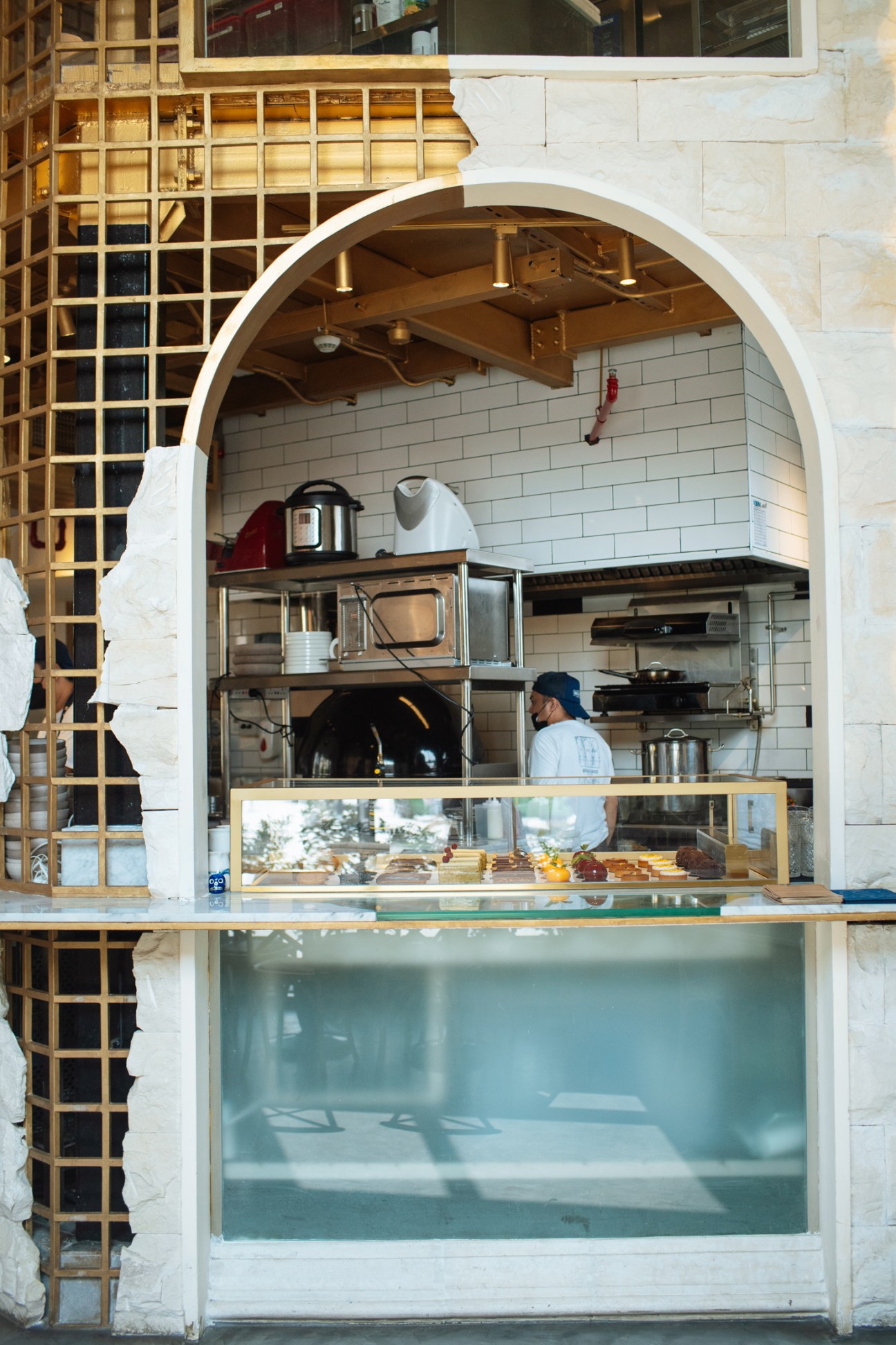 A modern café storefront with arched white entrance, displaying pastries behind glass counter with staff visible in kitchen area.
