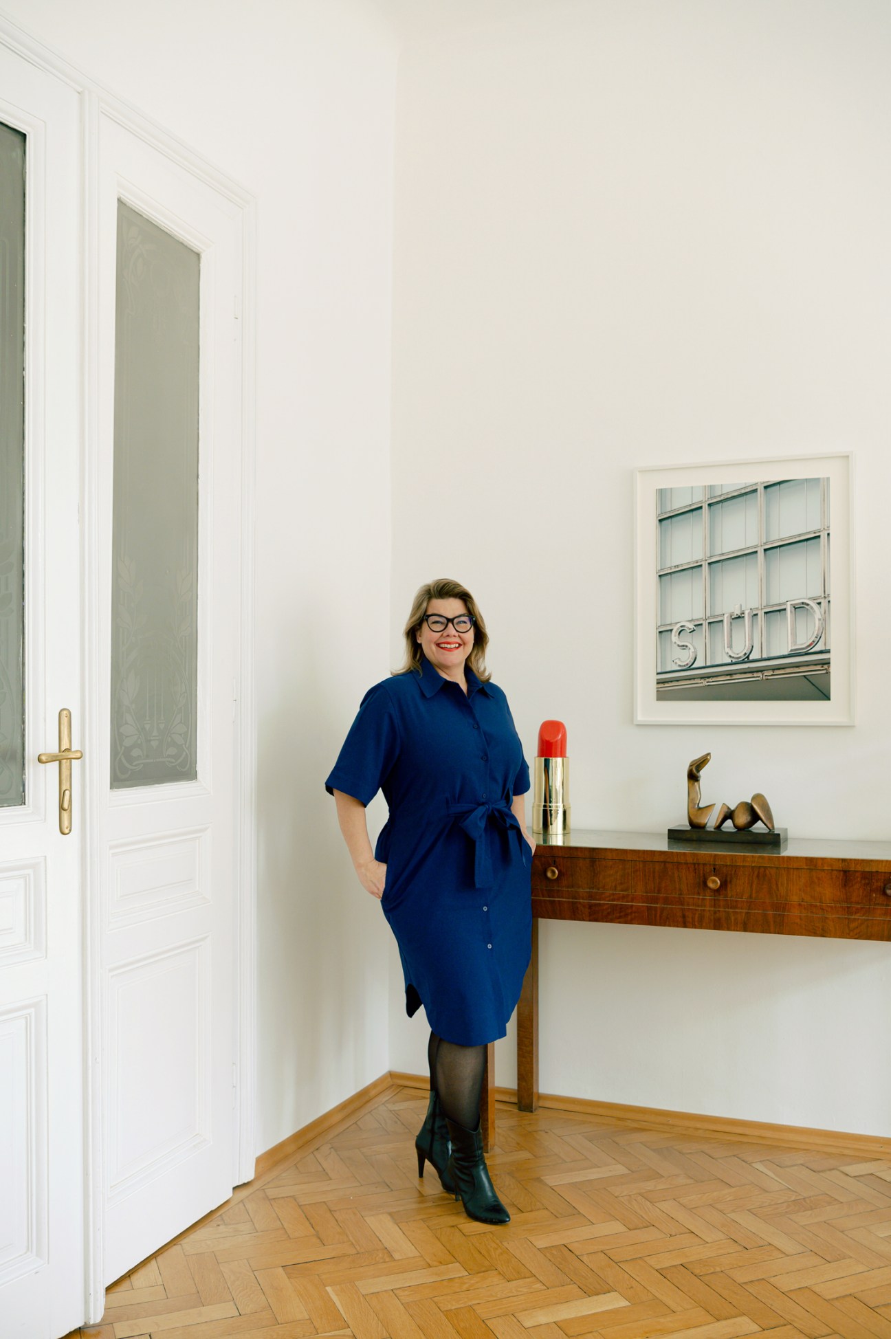 Woman in blue shirt dress standing in modern Vienna apartment with wooden floor and framed artwork.