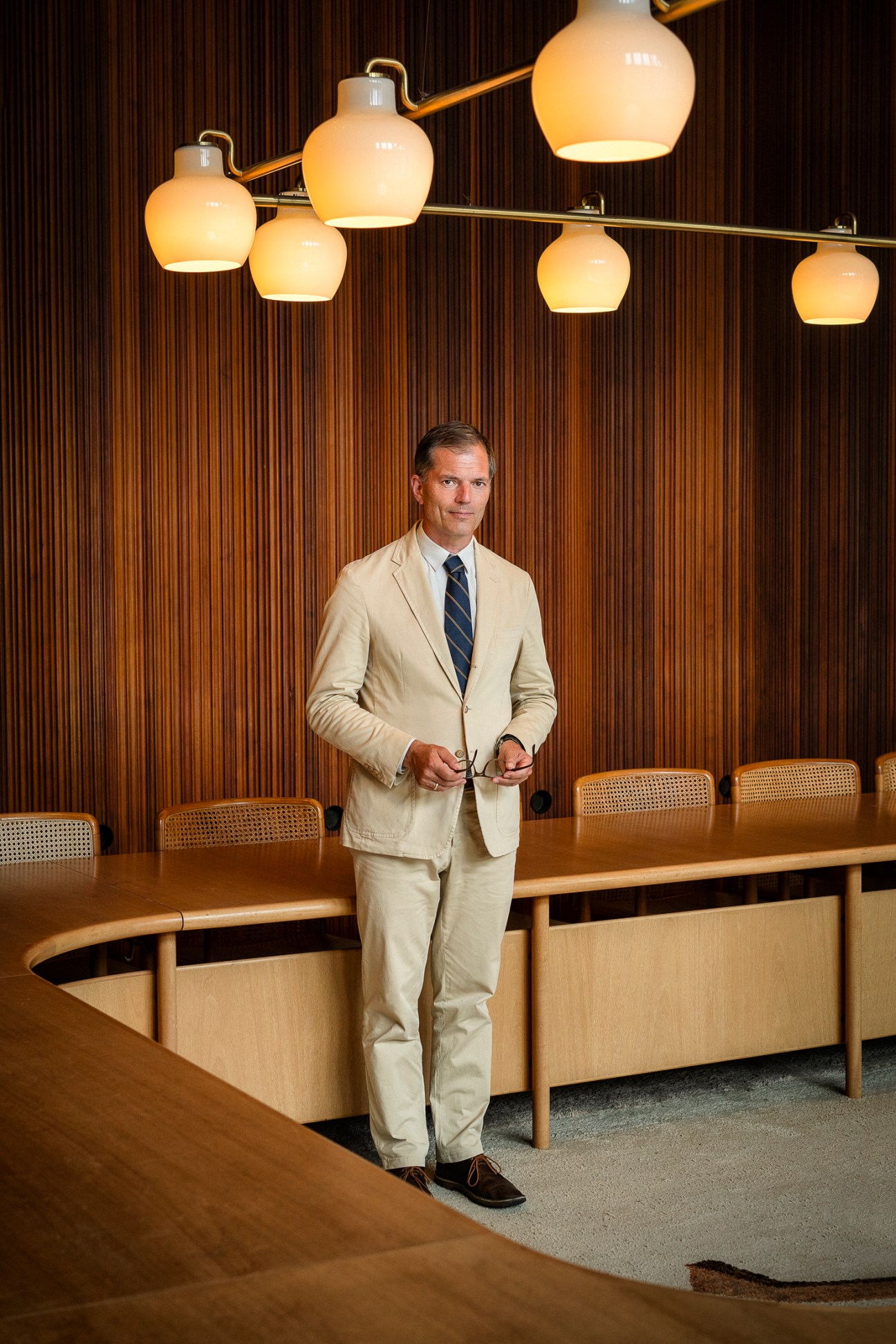 Man in beige suit and navy tie standing in modern office with wood paneling and pendant lights.