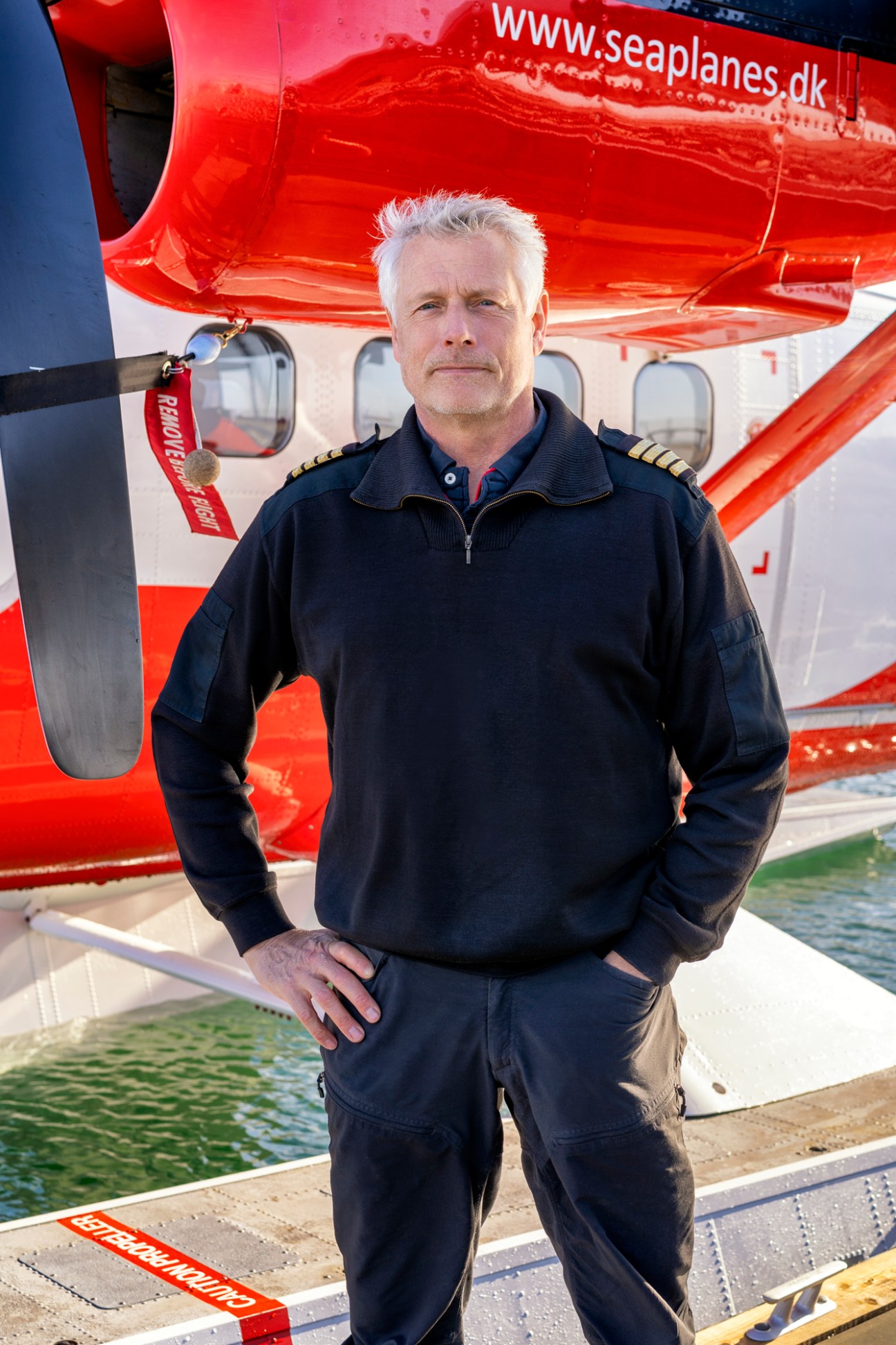 Seaplane captain in dark uniform with golden epaulettes standing beside a red Twin Otter aircraft at a waterside dock.