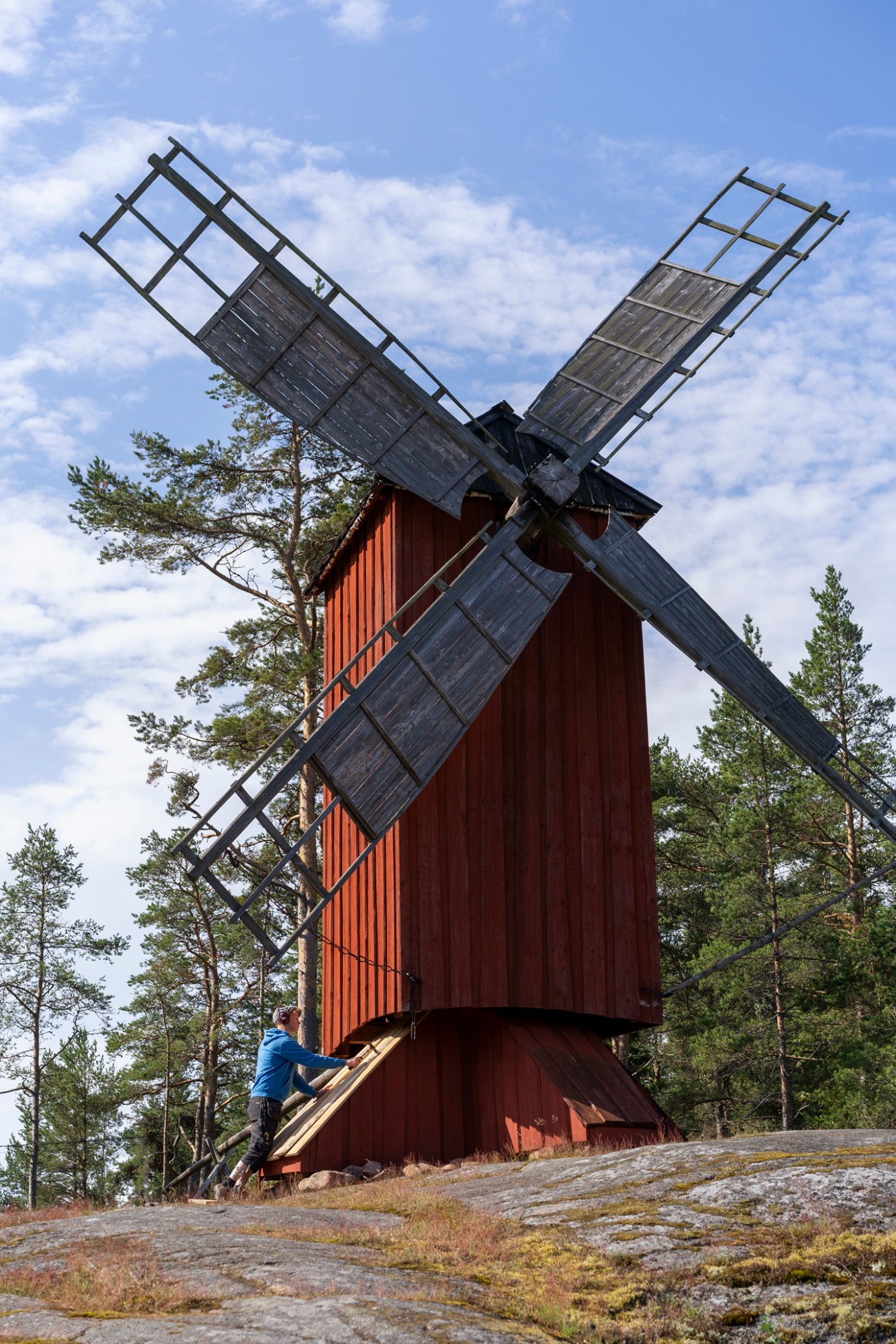 Traditional red wooden windmill with four wooden sails on rocky terrain surrounded by pine trees