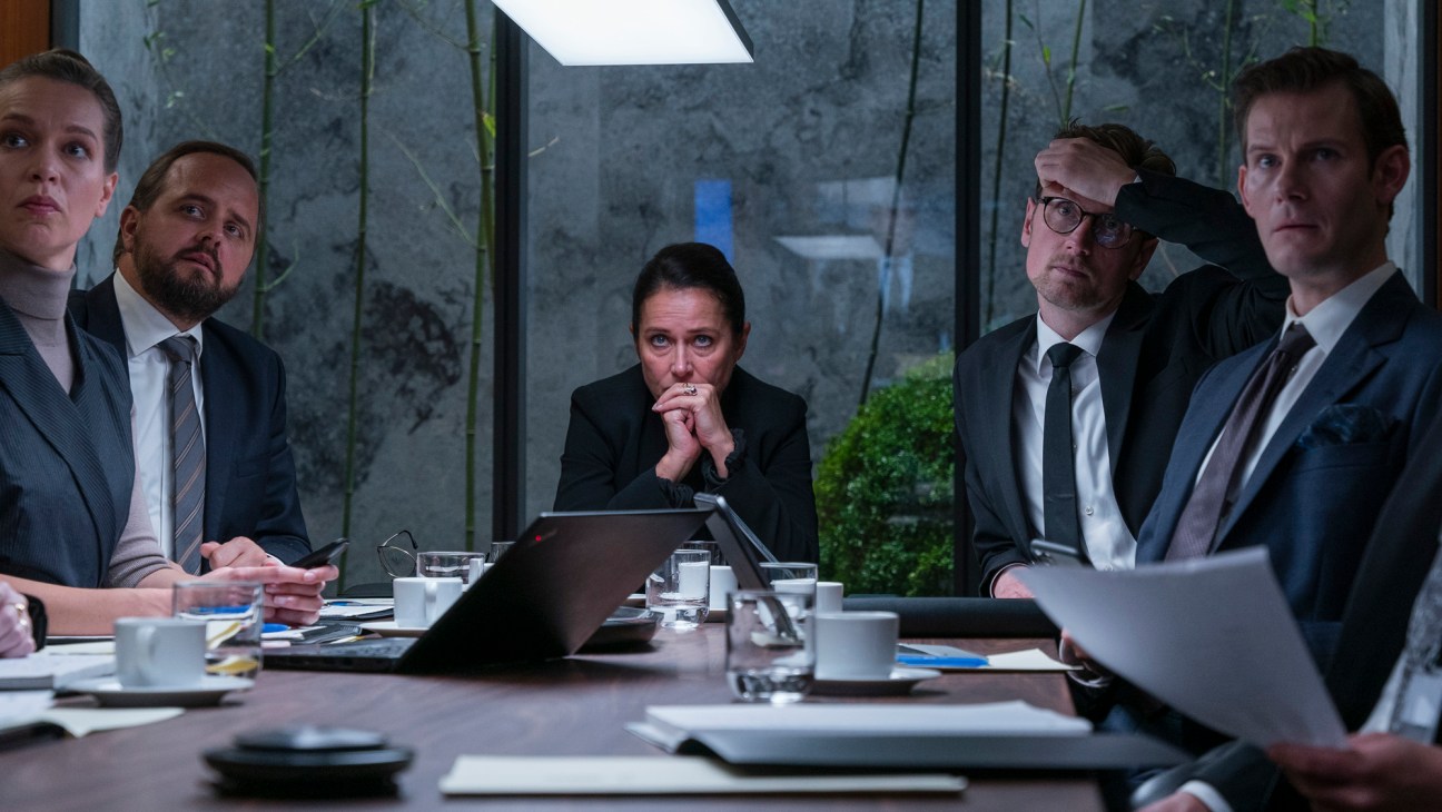 Five people in business attire sitting around conference table looking tense during meeting