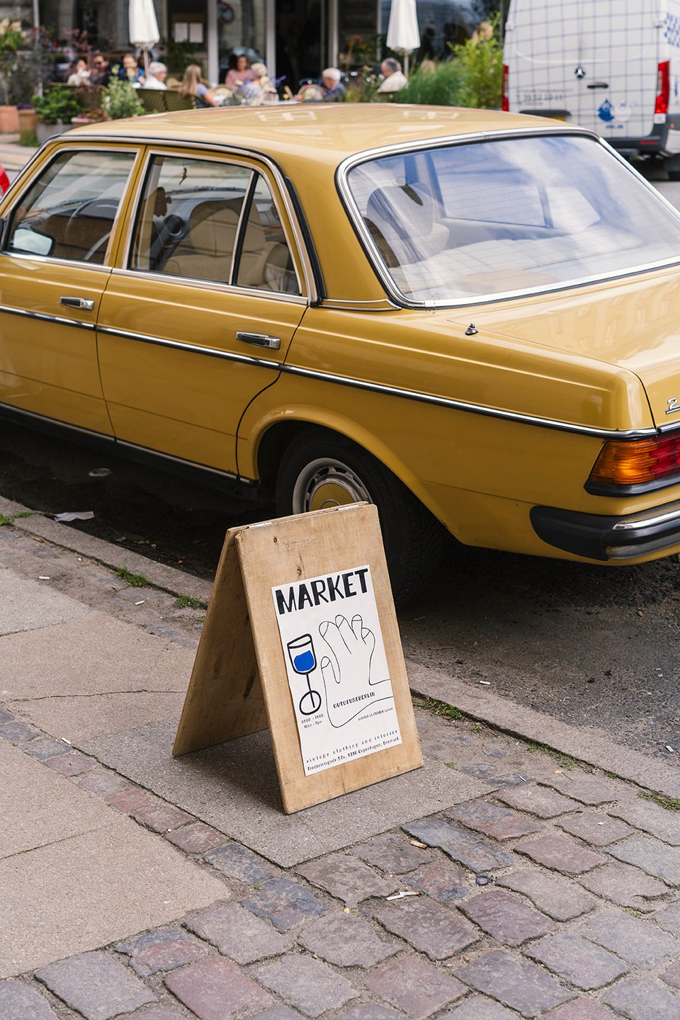 A simple brown wooden A-frame sign on a cobblestone street reading MARKET with a small blue illustration