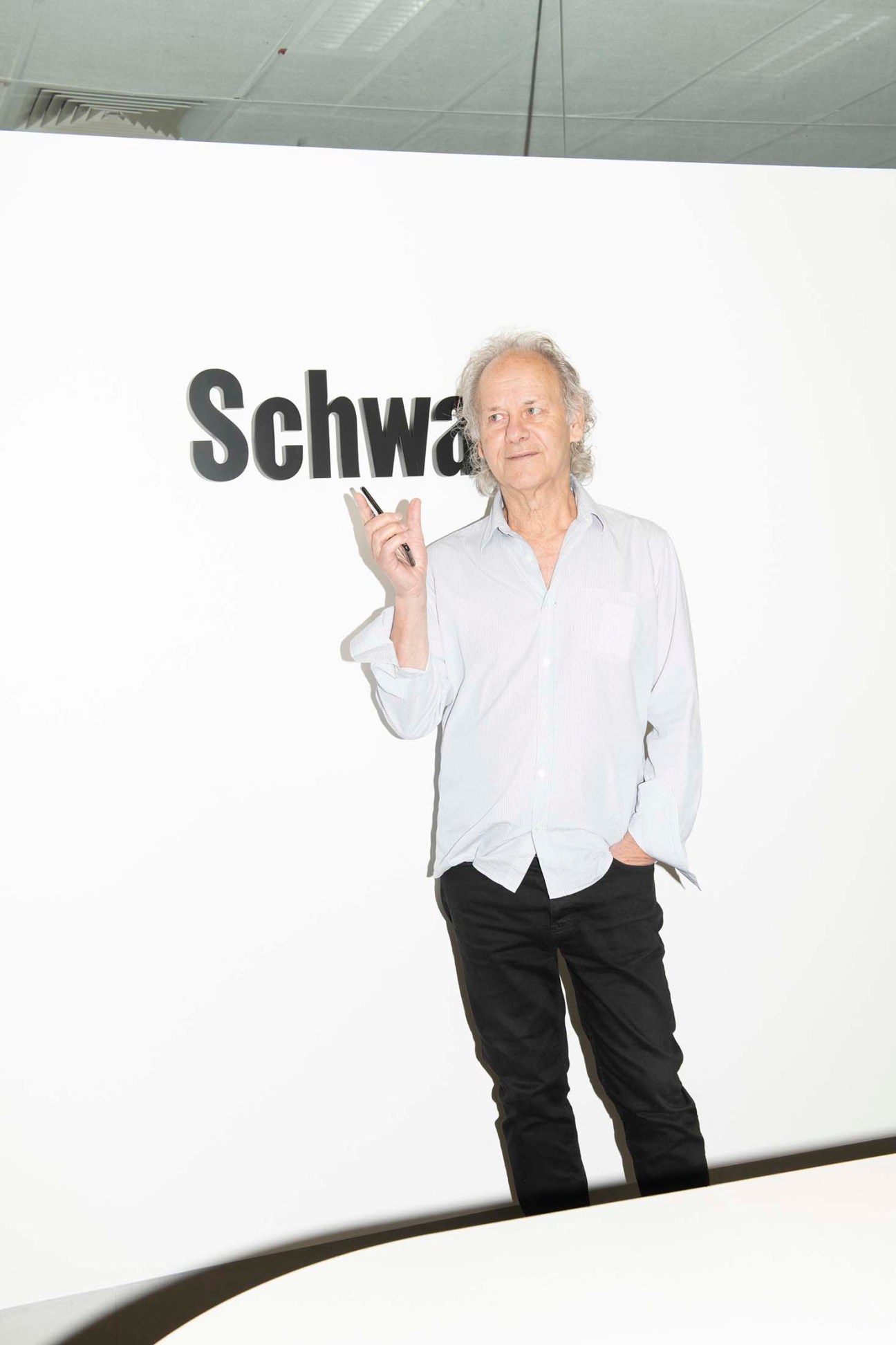 Morry Schwartz, an older man with gray hair wearing a white shirt and black pants, holds a cigar while standing in front of a white wall with partial text reading 'Schwa'