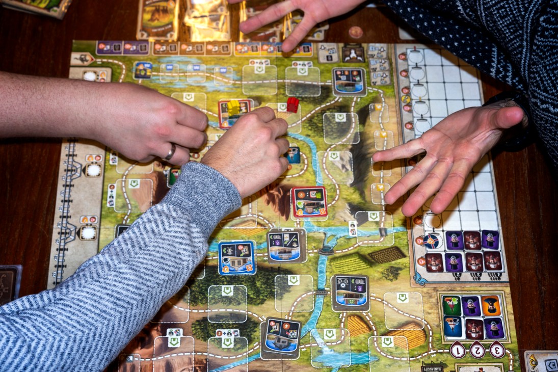 Two players' hands positioned over a complex board game with a green landscape board at Copenhagen's Board Games Club.