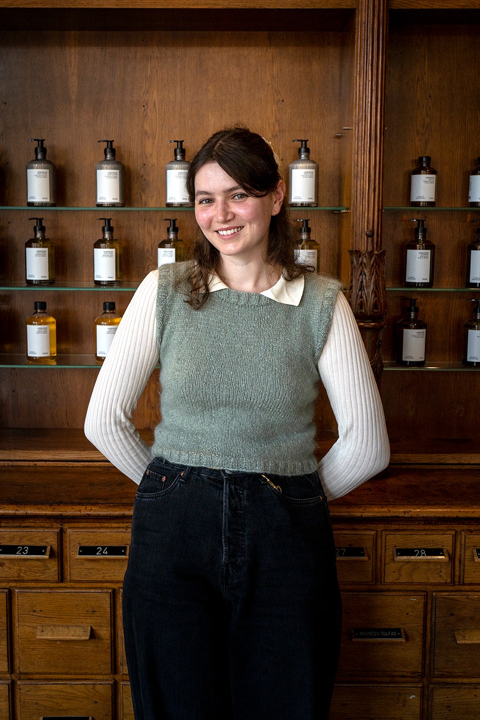 A smiling woman with brown hair wearing a gray sweater vest and white shirt standing in front of wooden shelves