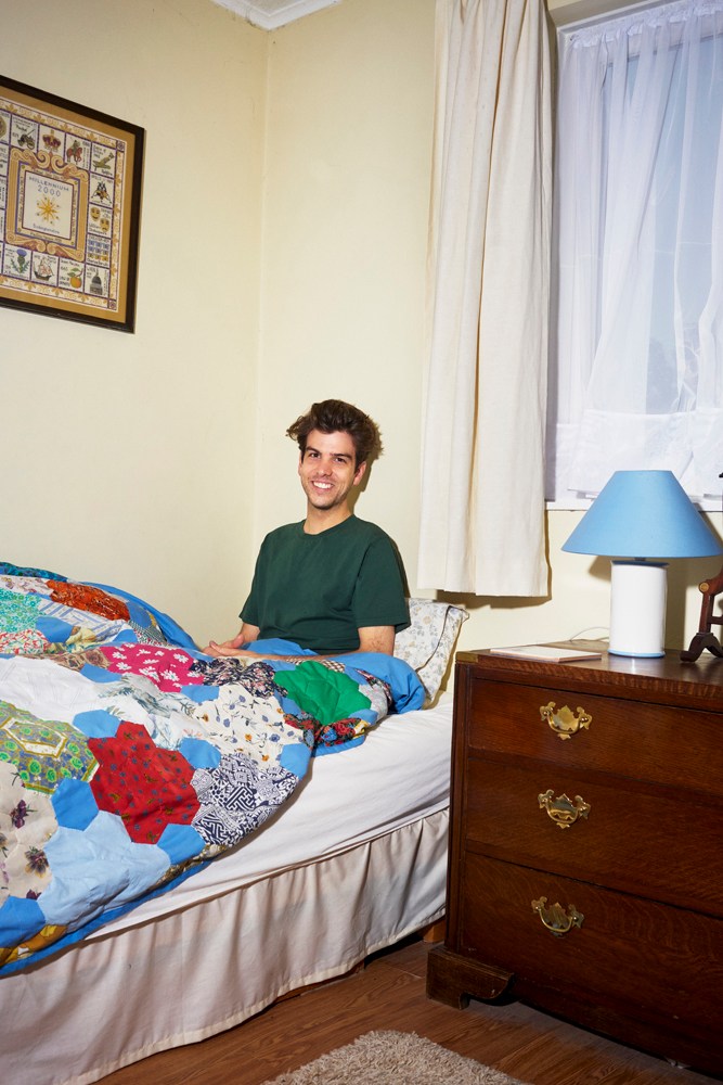 Man sitting on a bed with a colorful patchwork quilt in a modest bedroom with pale yellow walls and a blue lamp.