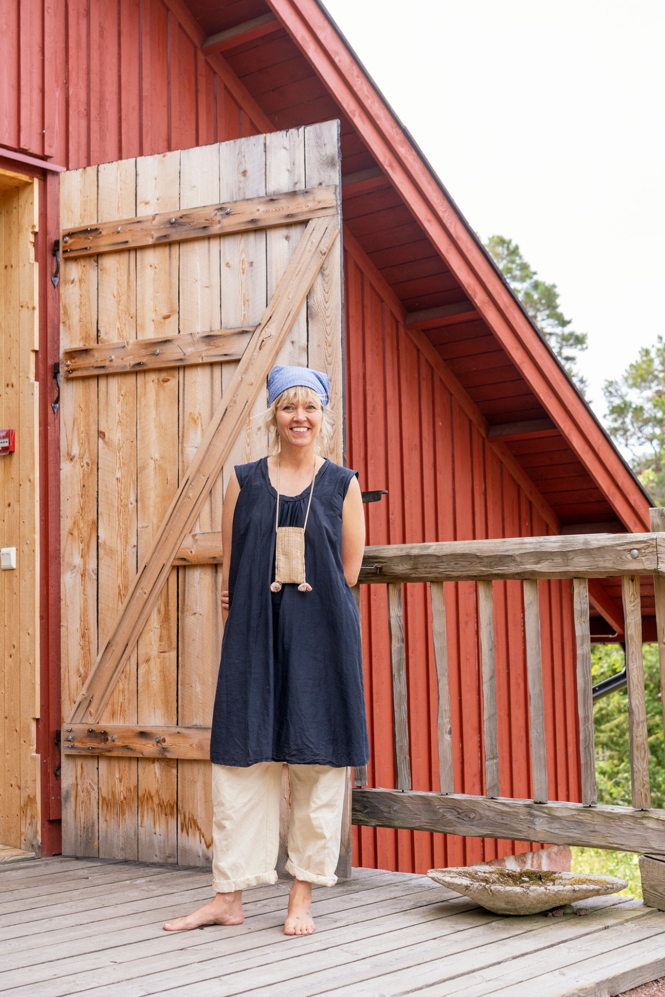 Lotta Jansdotter standing on wooden deck in front of red barn wearing navy dress and headband