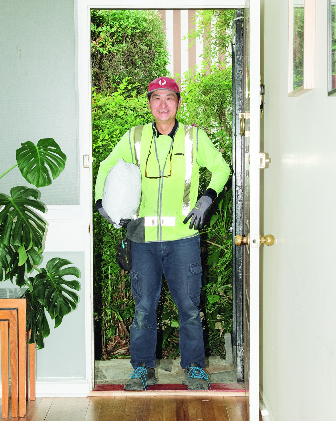 Delivery driver in hi-visibility vest and red cap holding white helmet, standing in residential doorway during pandemic isolation.