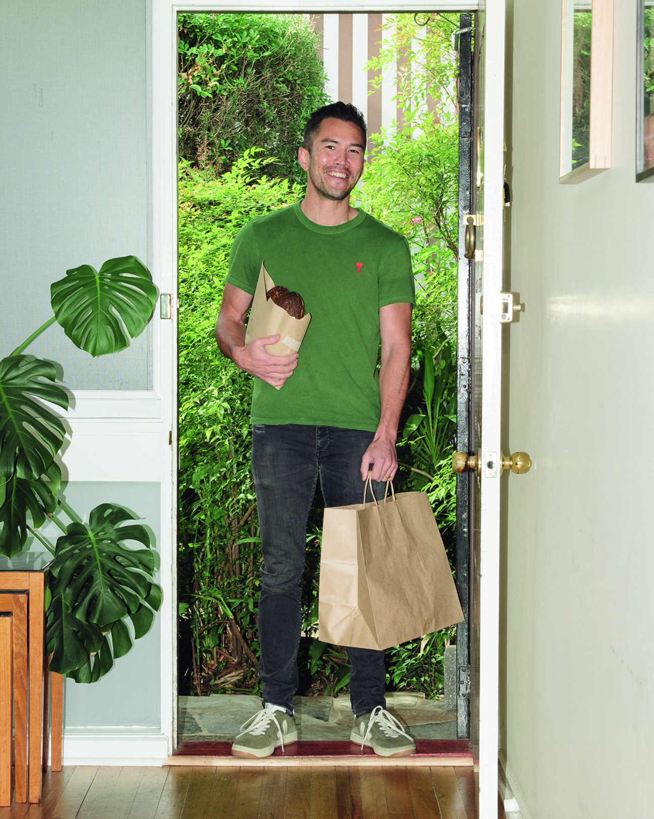 Man in green t-shirt holding food delivery bag at doorway with plants in background.