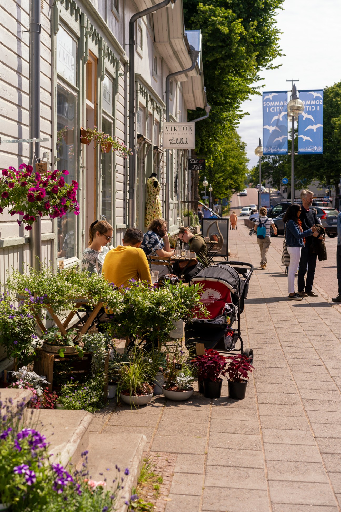 Outdoor cafe scene with people dining at tables along a pedestrian street with shops and flowering plants