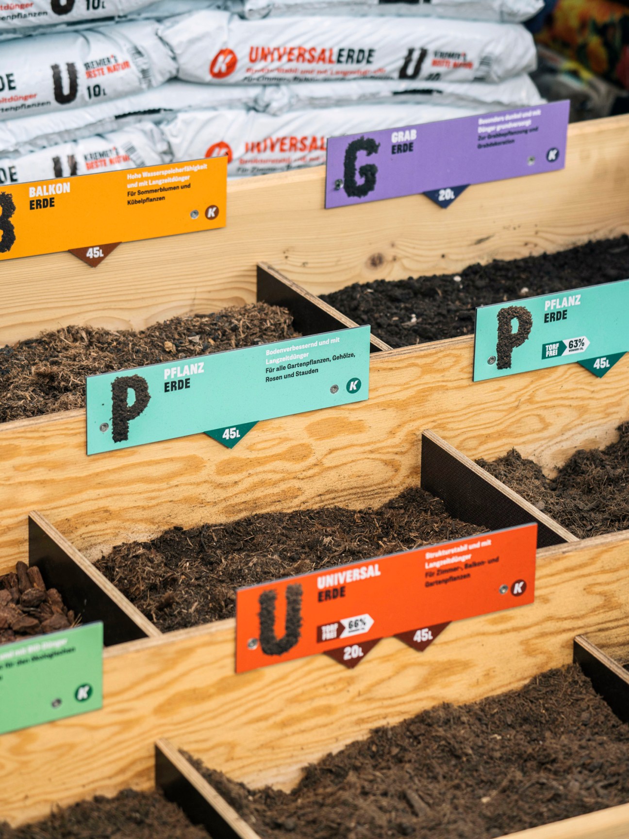 Wooden raised garden beds with colorful product labels for different soil types including Balkon Erde, Universalerde, Graberde, and Pflanzerde.