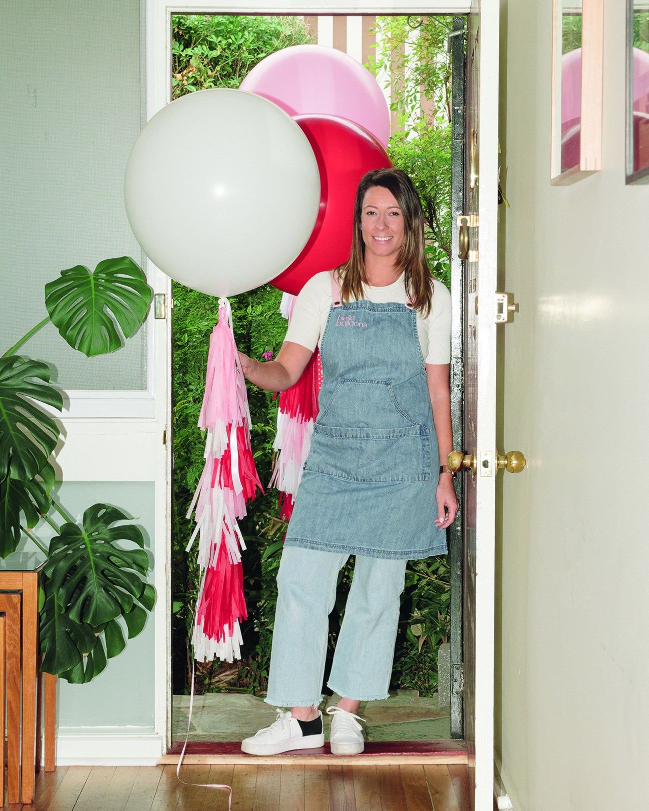 Woman in denim apron holding colorful balloons framed in her doorway during pandemic lockdown.