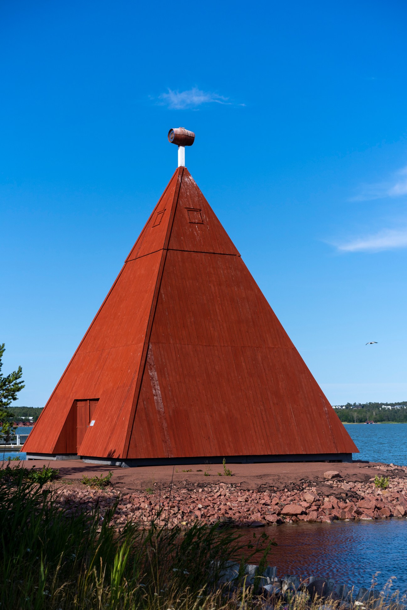 Large triangular red-brown wooden structure with white pole on top situated on waterfront with lake in background