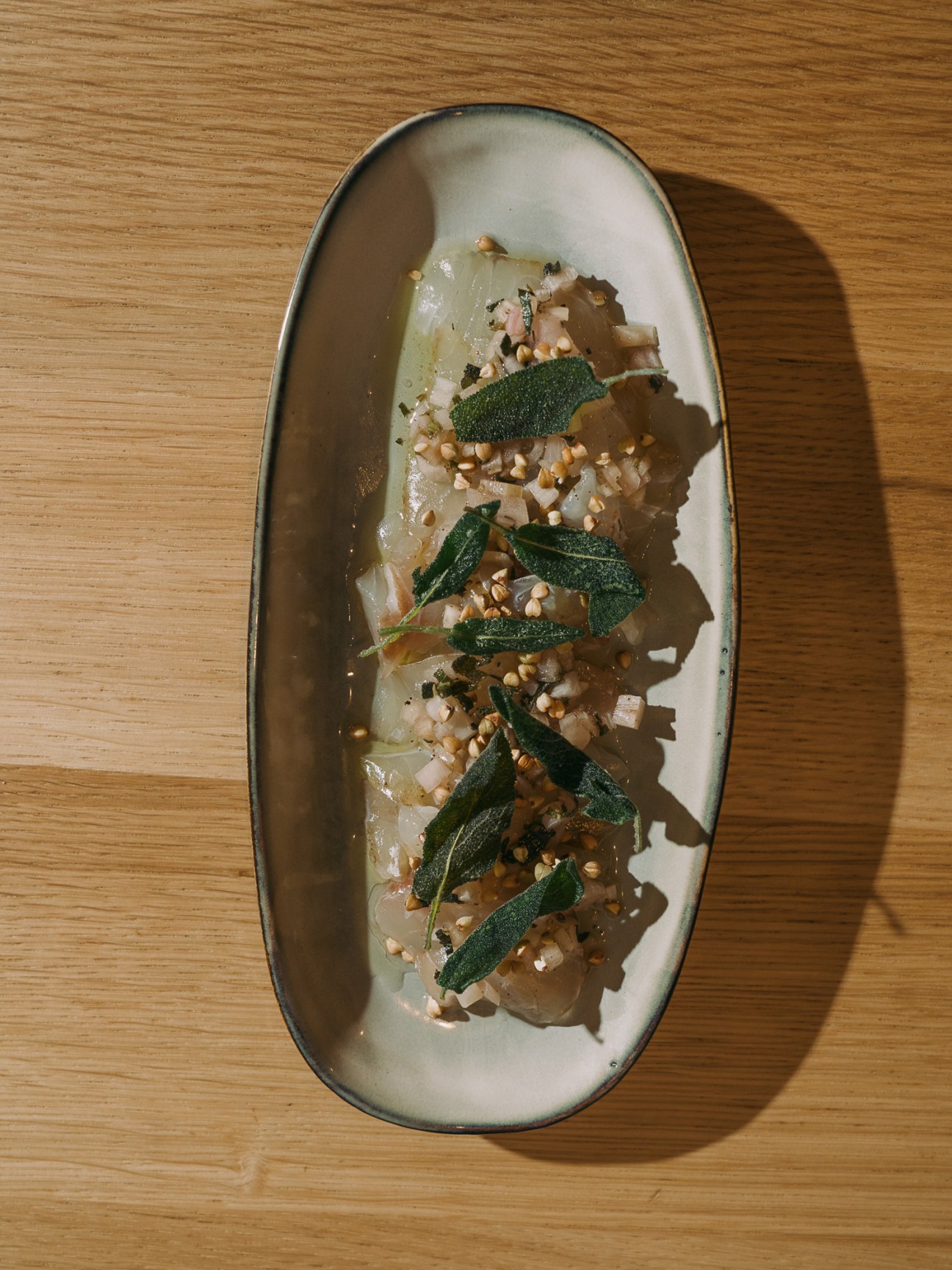 Sea bream carpaccio with fried sage leaves and chopped nuts on an oval ceramic plate