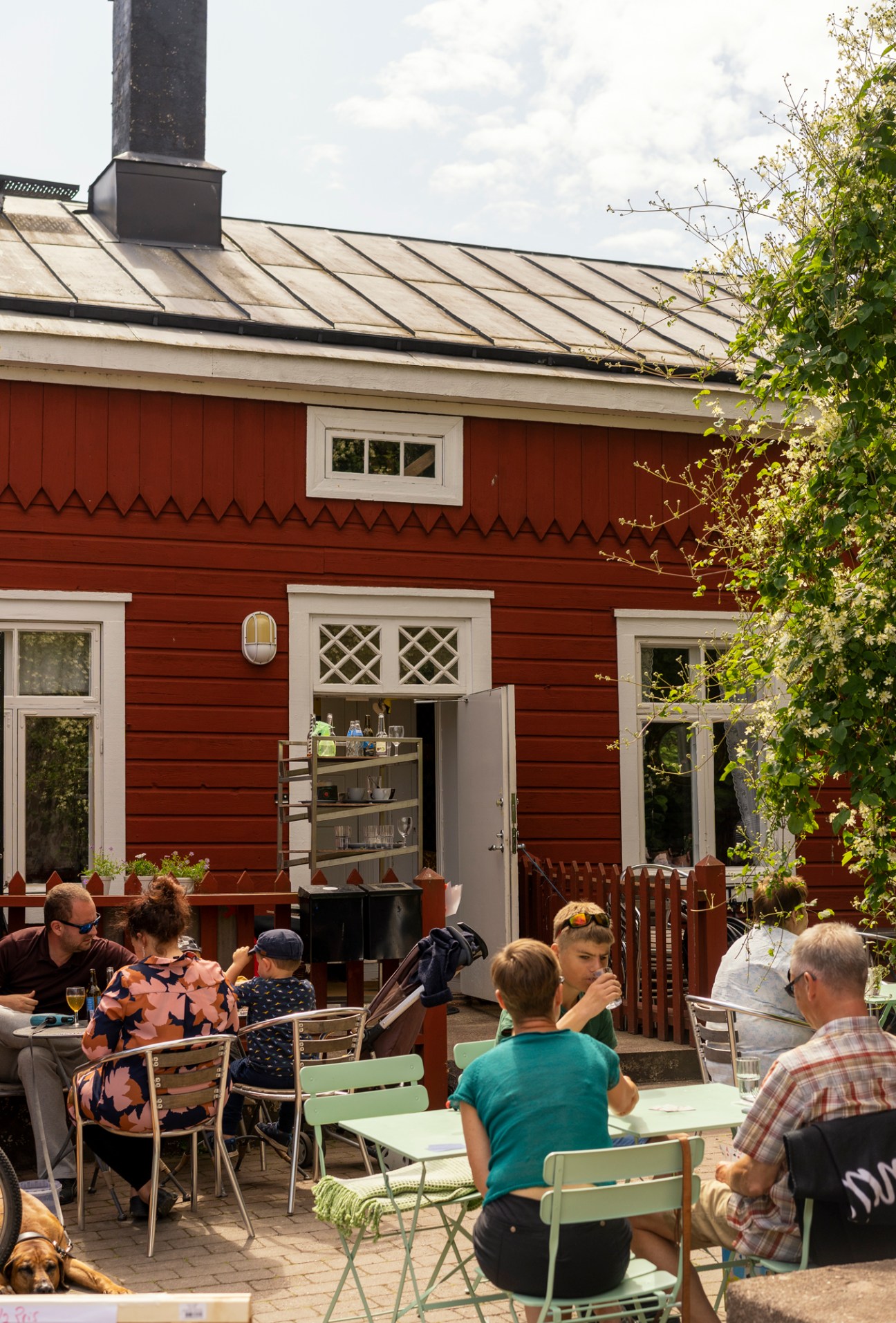 Red wooden building with white trim and outdoor seating area where people dine at tables on sunny day