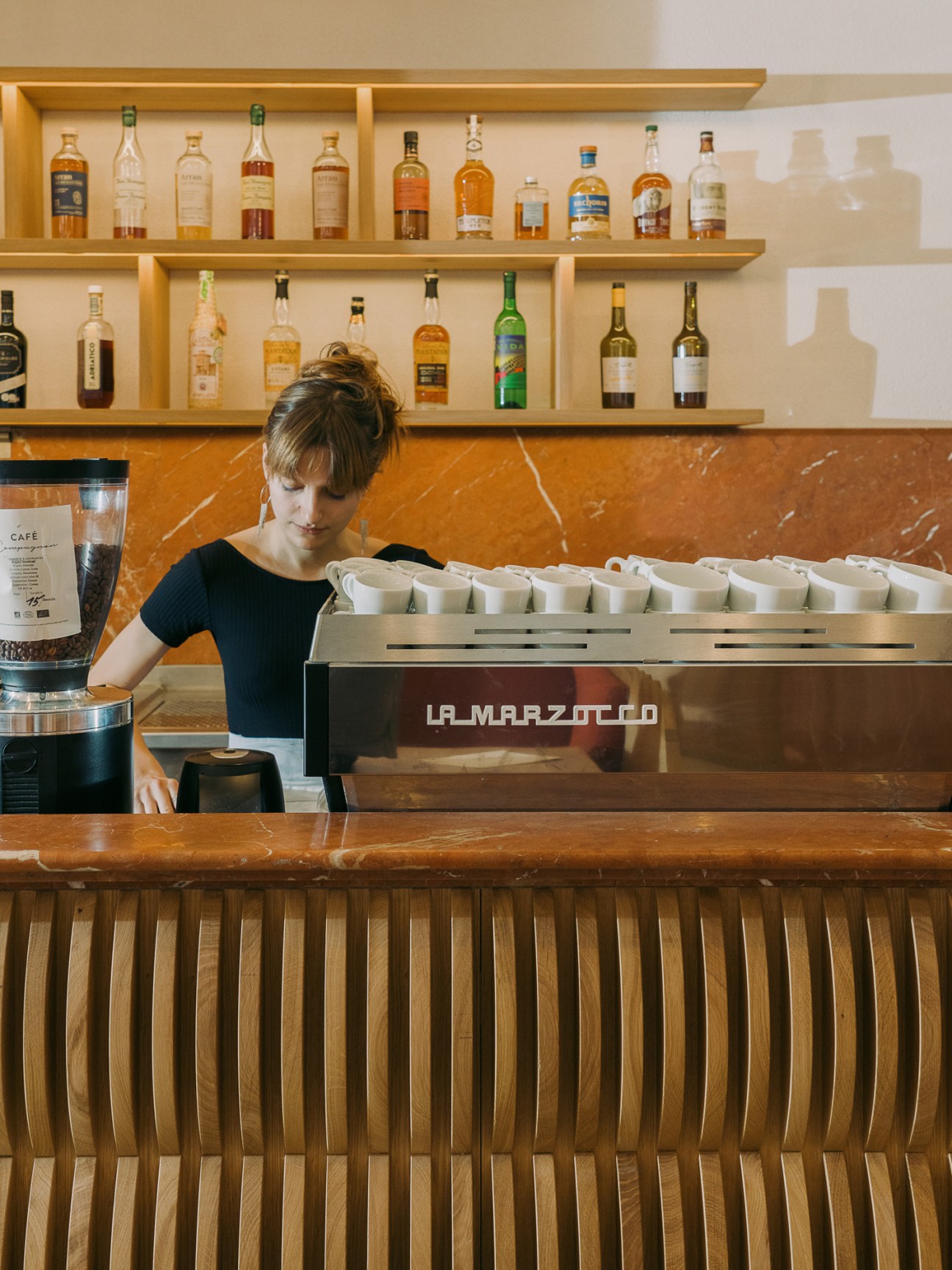 Barista working behind La Marzocco espresso machine with white coffee cups lined up on counter