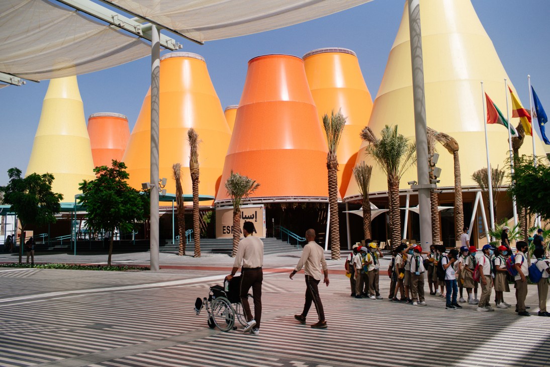 Large colorful cone-shaped buildings with yellow and orange fabric coverings in a plaza with palm trees and visitors.