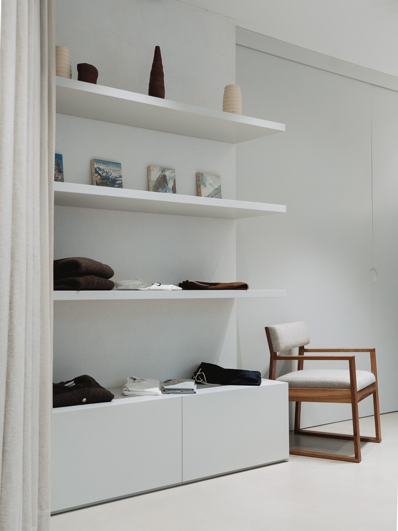 Interior of De Bonne Facture clothing store with white built-in shelving displaying folded garments and a wooden chair