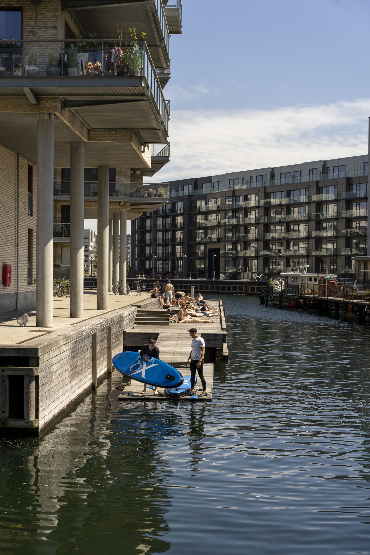 People with a blue paddleboard on a waterfront dock beside modern brick buildings.