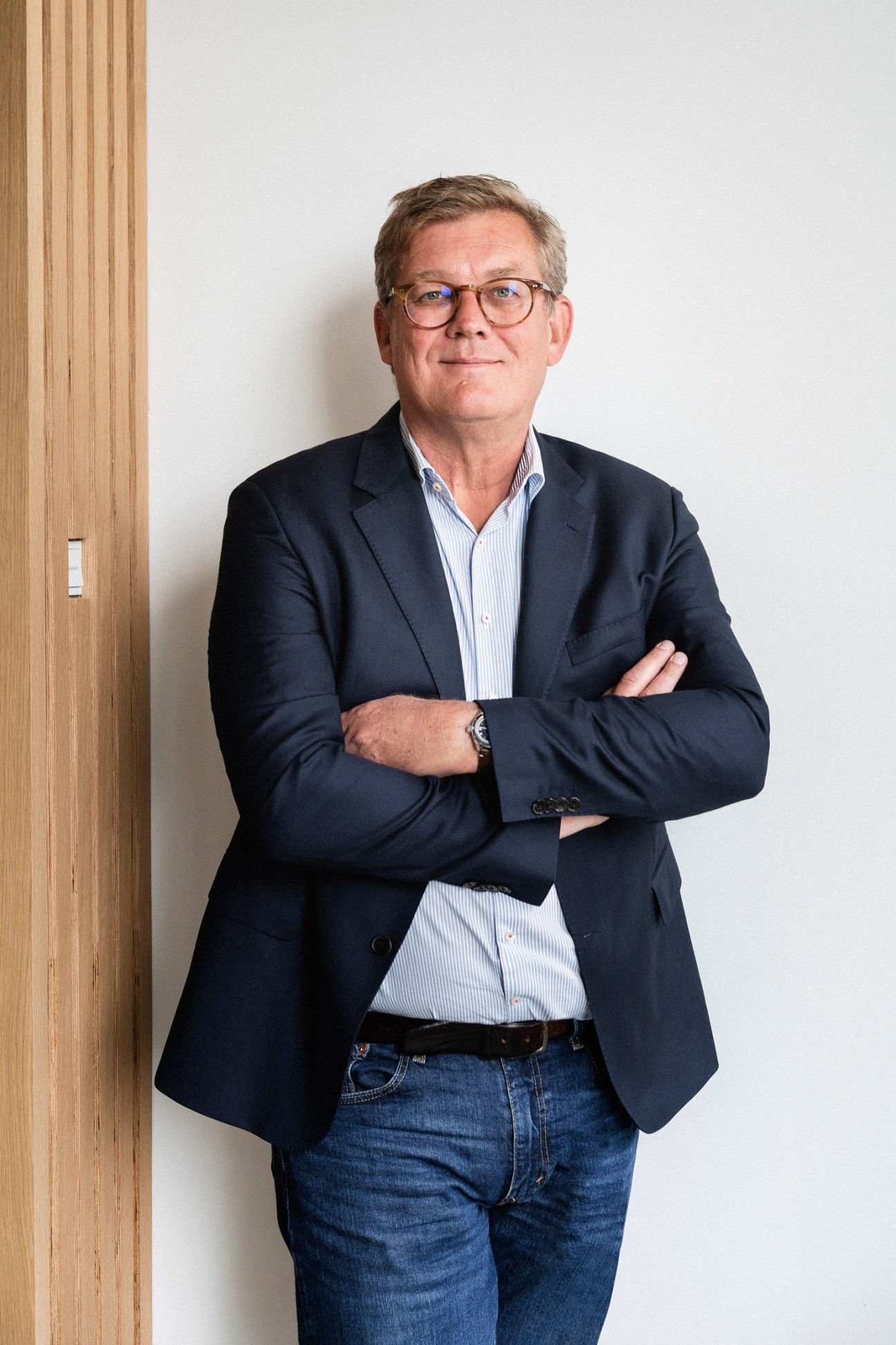 Jacob Nybroe stands with arms crossed wearing glasses, navy blazer and striped shirt in modern office space
