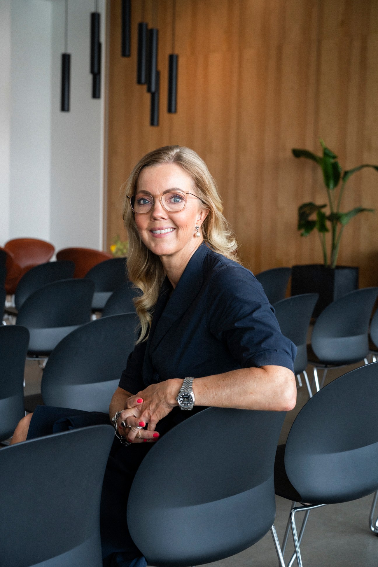 Trine Korsgaard Christensen, blonde woman with glasses and navy blazer, sitting in modern office with gray chairs