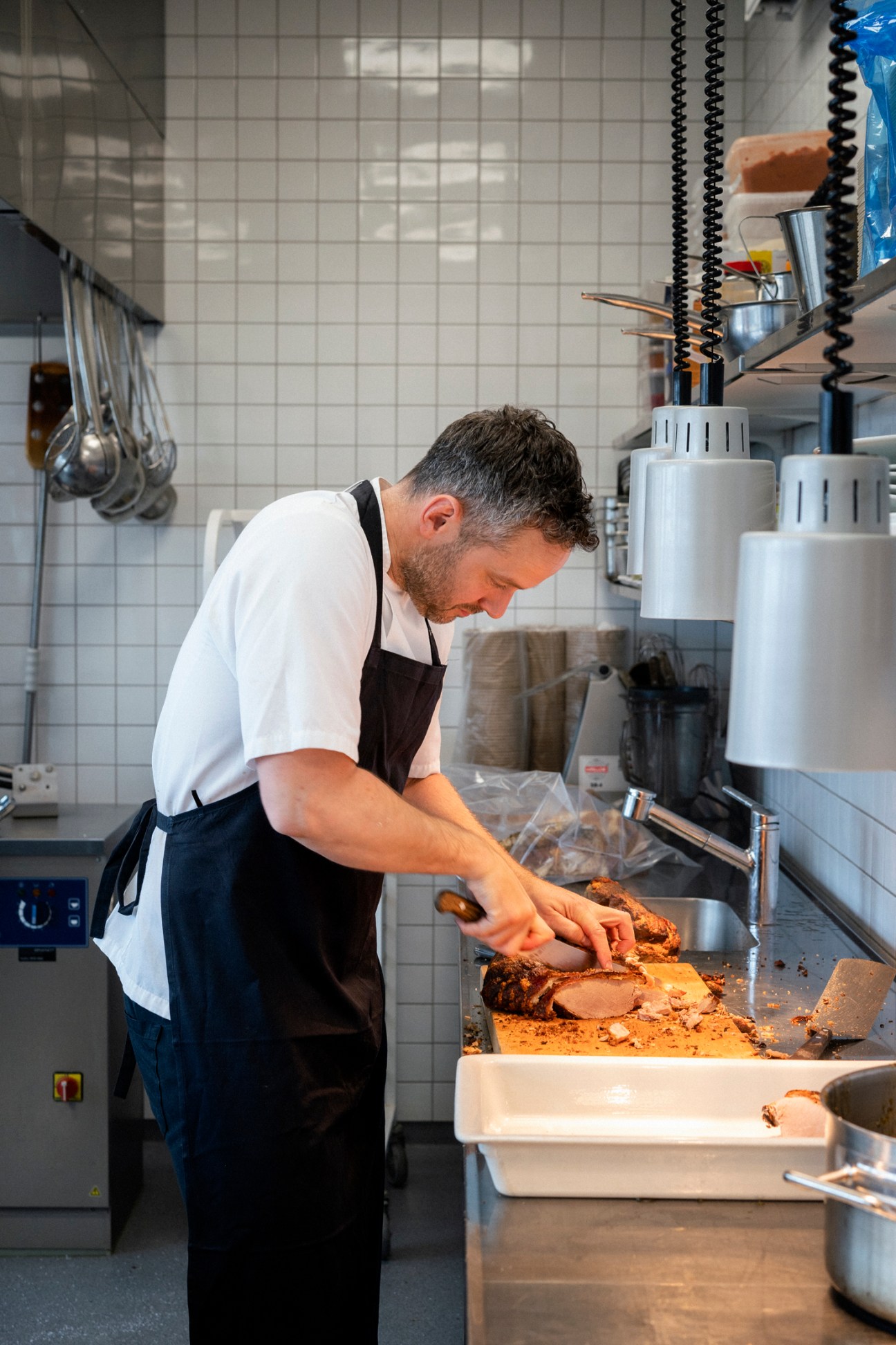 Chef in white shirt and black apron slicing roast pork in professional kitchen with white tiled walls