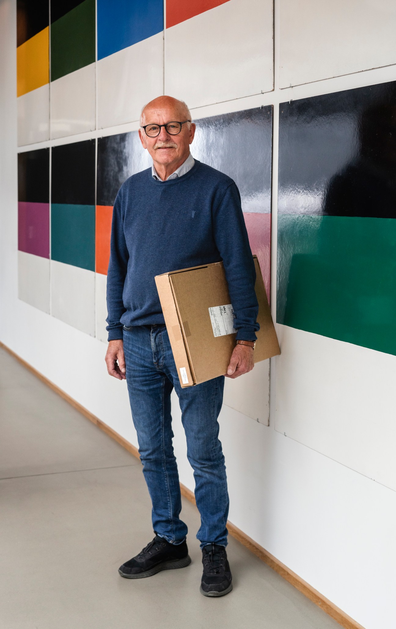 Older man with glasses and mustache holding cardboard package in modern office hallway with colorful wall art