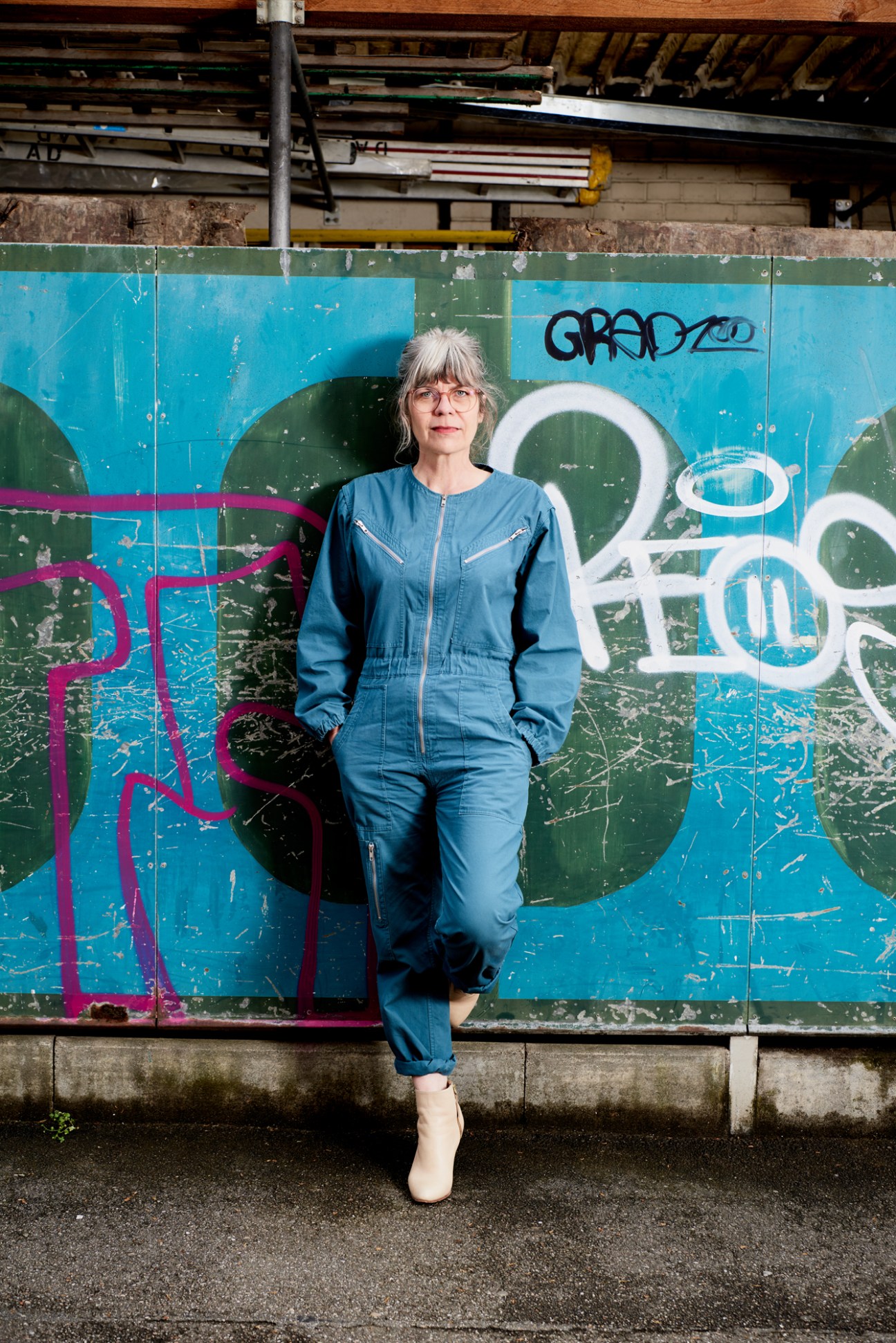 Woman in denim jacket leaning against colorful graffiti wall beneath industrial warehouse structure