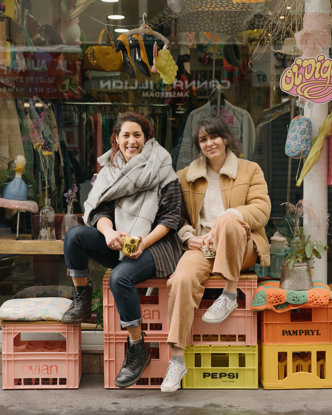 Two women smiling while sitting on colorful plastic crates outside a vintage clothing shop