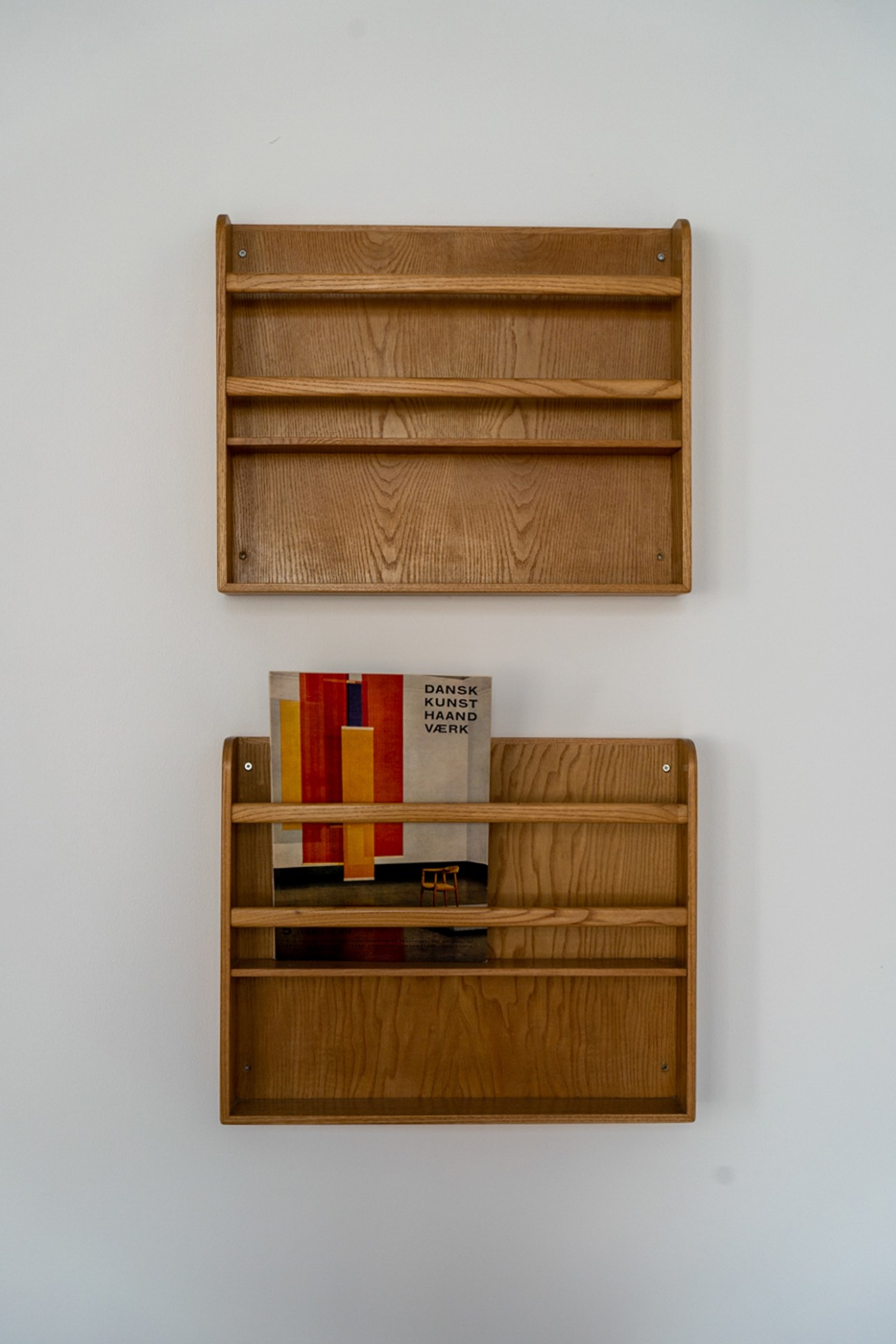 Two wooden wall-mounted book display shelves, one empty and one holding colorful books including Danish design publication