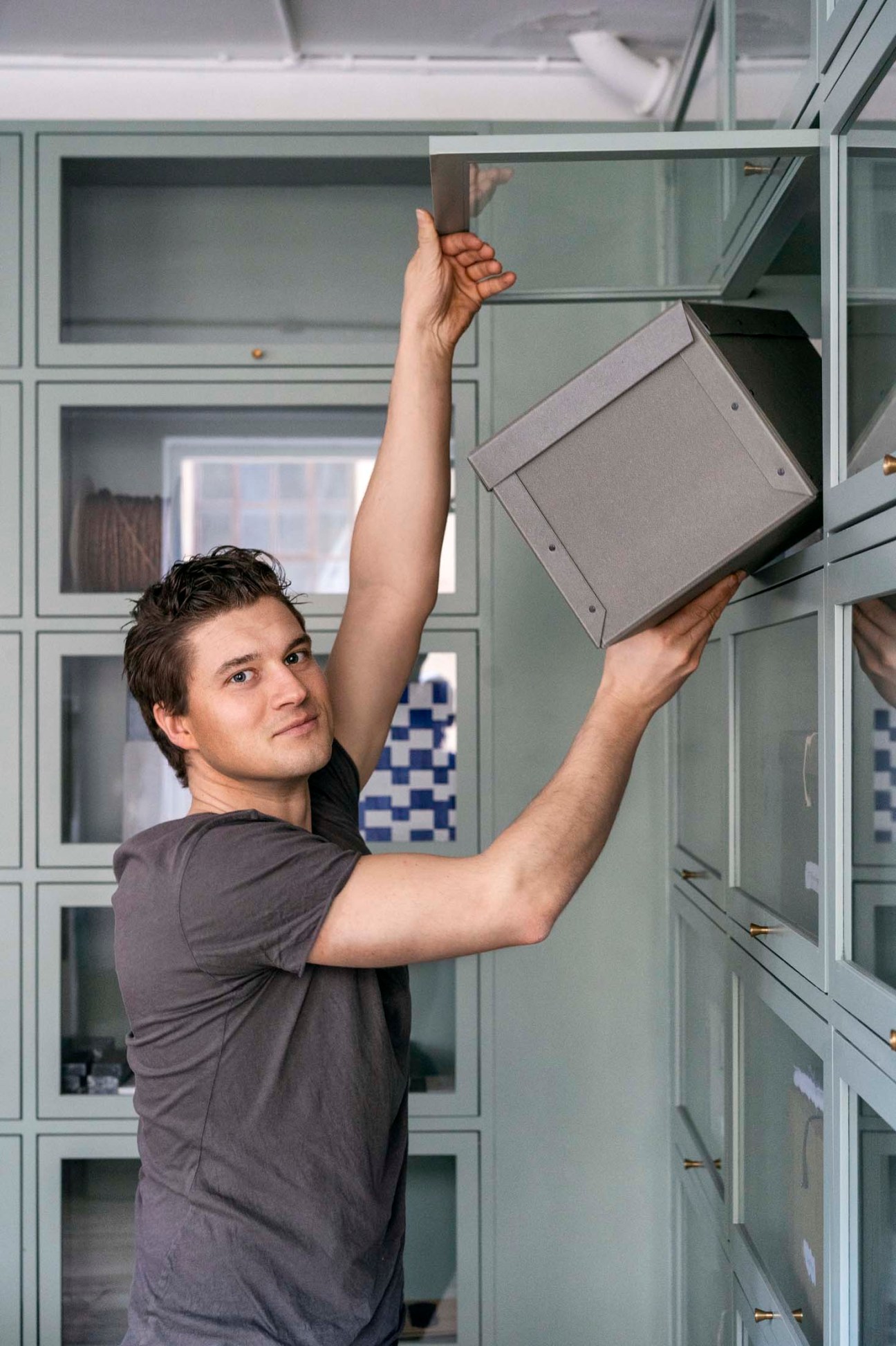 Man placing gray cardboard storage box on wall-mounted shelving system in bright interior space.
