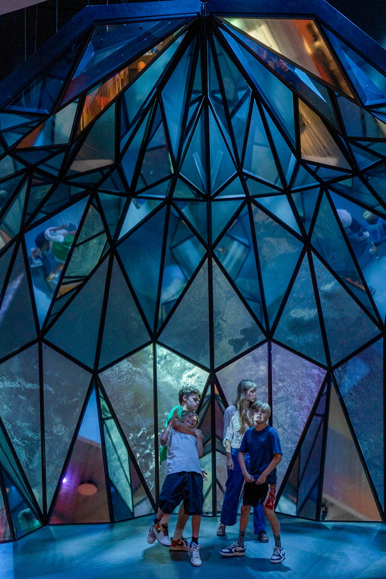 Children standing inside geometric glass dome structure with blue-tinted panels and metal framework
