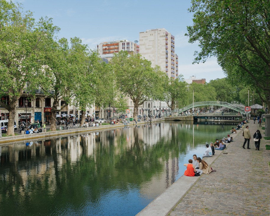People relaxing along the Canal Saint-Martin in Paris, with trees lining the waterfront and a bridge spanning the canal.