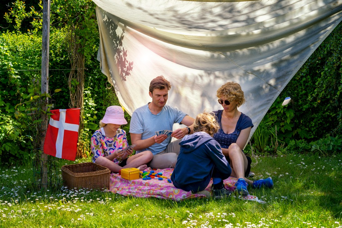 Family with children playing cards under white canopy at Danish allotment garden with flag