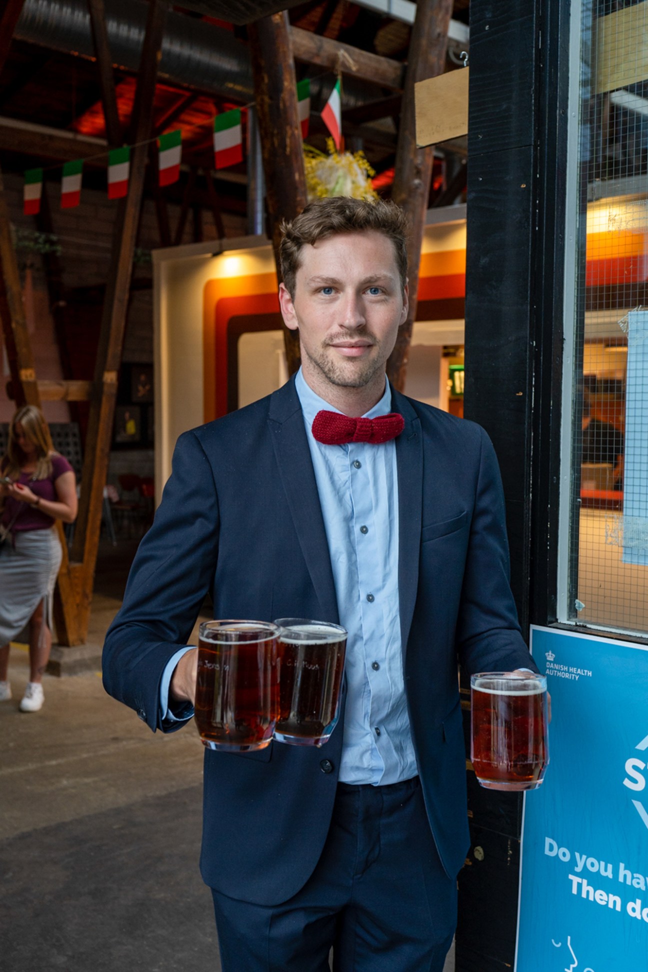 Man in navy suit and red bow tie holding three glasses of beer at Storms Pakhus